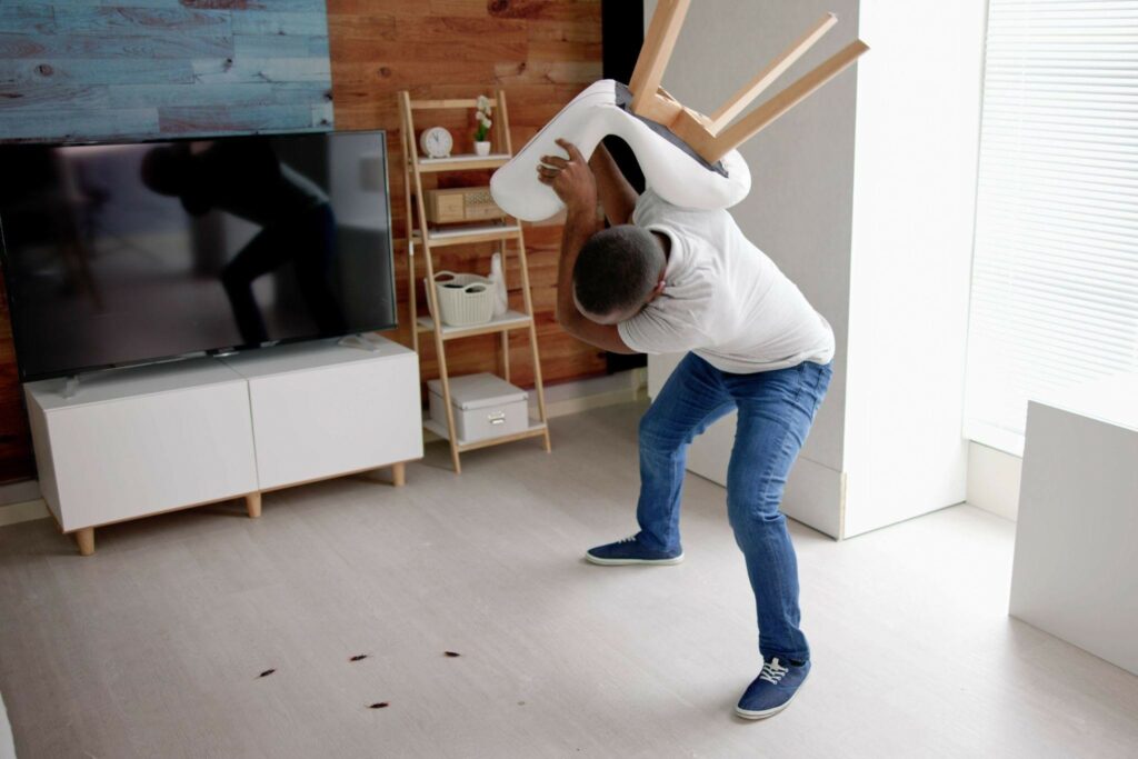 a black man in a white t shirt and blue jeans holding a chair above his head trying to smash bugs that are on the ground